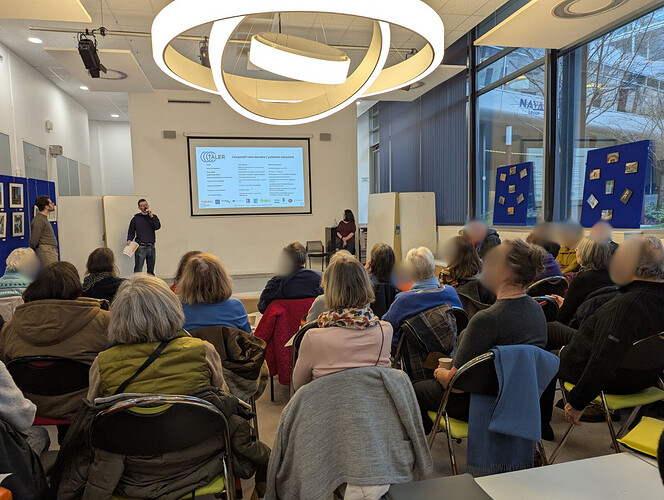 A community event organized by E-Seniors is taking place in a bright, modern room. Around 60 attendees, mostly seated on chairs, face a presentation area at the front. A speaker stands beside a projected slide introducing the NGI TALER project, explaining its outcomes, how it works, and the benefits of instant payments. Another person stands nearby, while a third sits to the side, ready to participate. Large windows line the right wall, letting in natural light, and display boards with photos are set up around the room. Overhead, a large circular light fixture hangs from the ceiling, illuminating the audience as they listen attentively to the presentation.